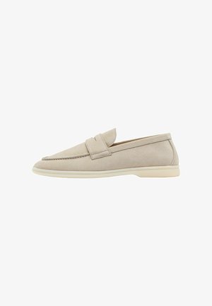 Zapato slip-on de ante beige con puntera redondeada, banda decorativa en la parte frontal y suela de goma ligera. Textura suave y diseño minimalista.