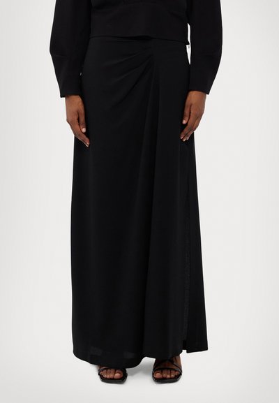 Schwarzer Maxikleid aus glattem Material, mit drapiertem Frontdetail, Schlitz an der Seite und fließender Silhouette. Kombiniert mit schwarzen Sandalen mit Riemchen.