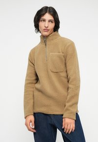 Tan fleece pullover met een hoge kraag, halve rits en een borstzak. Zachte textuur en een ontspannen pasvorm, gecombineerd met donkere jeans.
