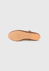 Une chaussure plate brune à clous, posée semelle vers le haut sur un fond blanc, montrant la semelle texturée et le nom de la marque "Steve Madden".