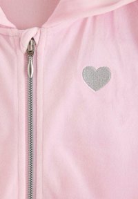 Lyserød fleece zip-up hoodie med en sølvfarvet hjertebrodering på brystet. Har en metal lynlås og en blød tekstur.