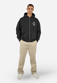 Černá mikina na zip s grafickým potiskem, předními kapsami a žebrovanými manžetami, spárovaná se béžovými tepláky a bílými teniskami.