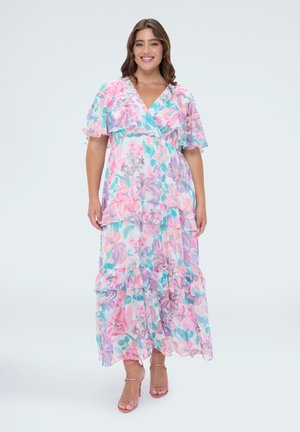 Lächelnde Frau in einem pastellfarbenen, geblümten Maxikleid mit Rüschenärmeln und Stufenrock, die in rosa Riemchen-High Heels steht.