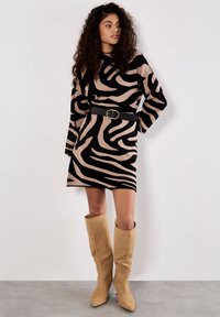 Vrouw met lang, krullend haar draagt een zwart-beige jurk met zebra-print, versierd met een zwarte ceintuur en knielange tan suède laarzen, staande tegen een witte muur.