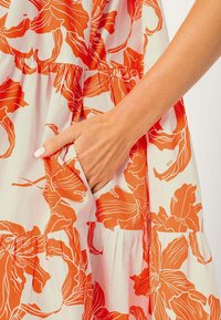 Högerhand med vit nagellack som gled ner i en ficka på en vit klänning med stora orange blommönster.