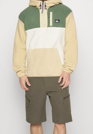 Personne portant un sweat à capuche polaire vert, blanc et beige avec une poche avant et un short cargo vert olive avec des poches zippées.