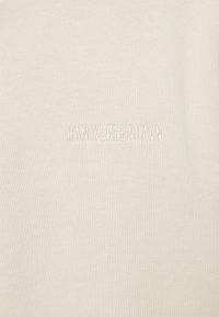 Ljusbeige t-shirt tillverkad av mjukt tyg, med subtil texturerad broderi av "HAN KJØBENHAVN" nära botten. Enkelt design.