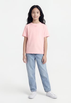 Κοπέλα με μακριά σκουρόχρωμα μαλλιά φοράει ανοιχτό ροζ μπλουζάκι, ανοιχτά μπλε τζιν και λευκά αθλητικά παπούτσια, στέκεται μπροστά σε απλό λευκό φόντο.