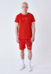 Conjunto de camiseta roja y pantalones cortos, que presenta el logo "Reject x Paris" en dorado, con mangas cortas, un corte relajado y calzado blanco.