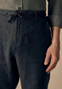 Pantalons en lin foncé avec taille à cordon de serrage, présentant une texture lisse et un léger éclat. Le gros plan montre une main reposant sur le tissu.