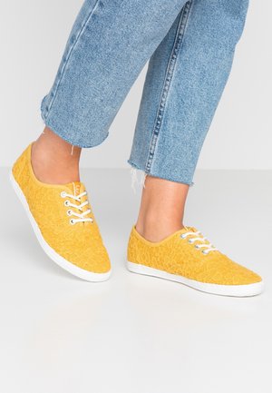Baskets en dentelle jaune avec semelles en caoutchouc blanc et lacets. Une texture en dentelle décorative recouvre le dessus, avec un bout rond et un design minimal.