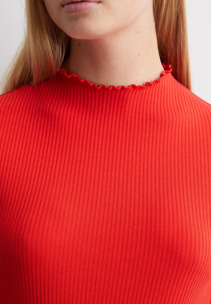 Rotes geripptes Oberteil mit hohem Kragen und gewelltem Saum. Die Textur ist glatt mit schmalen vertikalen Linien und passt eng am Körper.