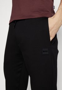 Black jogger pants with a smooth texture, featuring a side pocket and a square black logo patch labeled "BOSS." Paired with a burgundy top.