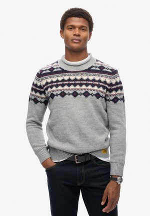 Suéter gris con cuello alto, que presenta un canesú estampado en azul marino, crema y burdeos. Se lleva con jeans oscuros y un reloj.