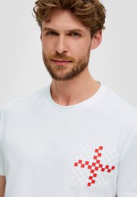 Camiseta de algodón blanca con un patrón de tablero de ajedrez rojo y blanco en la zona del lado izquierdo del pecho. Cuello clásico de tipo crew y mangas cortas.