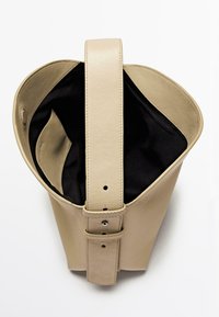 Béžová kožená kabelka se strukturovaným tvarem, horním uchem a černou vnitřní podšívkou. Má viditelné prošívání a kovový dekorativní prvek.