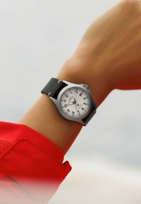 Montre en argent avec cadran blanc, chiffres arabes noirs et accents rouges. Équipée d'un bracelet en caoutchouc noir et d'un boîtier en acier inoxydable.