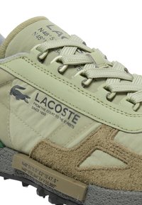 Primer plano de una zapatilla beige de Lacoste con detalles en ante texturizado, cordones tejidos y coordenadas impresas en la suela y la lengüeta.