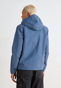 Giacca con cappuccio blu chiaro dal tessuto morbido, con polsini elasticizzati e orlo allungato, abbinata a pantaloni grigi scuri dotati di tasche.