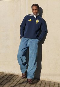 Námořnická mikina s švédským znakem, světlé modré volné džínové kalhoty a hnědé tenisky. Model se opírá o hladkou světle zbarvenou zeď.