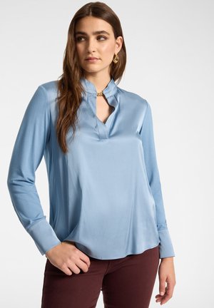 Femme aux longs cheveux bruns portant un chemisier en satin bleu clair et un pantalon foncé, regardant sur le côté avec une main posée sur la hanche.