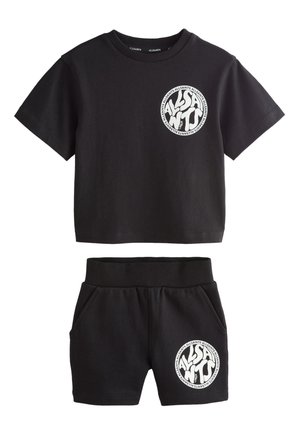 Junger Junge steht, trägt ein schwarzes passendes T-Shirt und Shorts mit weißen runden Logos, weiße Socken und weiße Sneaker.