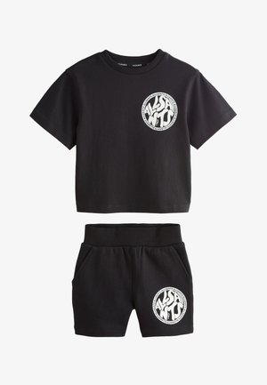 Junger Junge steht, trägt ein schwarzes passendes T-Shirt und Shorts mit weißen runden Logos, weiße Socken und weiße Sneaker.