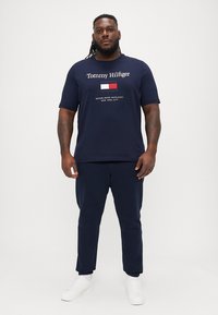 Marineblå bomulls T-skjorte med hvit "Tommy Hilfiger" logo og rød, hvit, blå emblem. Kombinert med marineblå joggebukser og hvite joggesko.