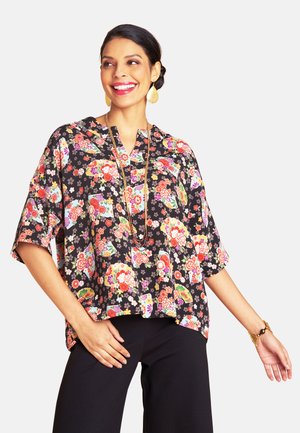 Florale Bluse aus schwarzem Stoff, mit kurzen Ärmeln, V-Ausschnitt und bunten Blumenmustern. Kombiniert mit dunkler Hose und einer Kettenhalskette.
