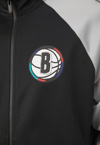 Μαύρο αθλητικό σακάκι με γκρίζες λεπτομέρειες, που διαθέτει κυκλικό λογότυπο με ένα μπάσκετ και το γράμμα "B". Κλείσιμο με φερμουάρ και υφή υφάσματος.