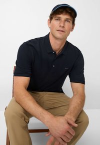 Navy blue polo shirt with a collar, two buttons, and a subtle logo. Worn with beige pants, seated posture, and light brown hair.