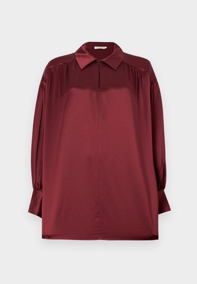 Langærmet bluse i bordeaux satin med spidst krave, plisserede skuldre og manchetter med knapper mod en ensfarvet baggrund.