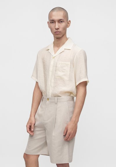 Homme portant une chemise beige clair à manches courtes avec poche avant et un short assorti jusqu’au genou, sur un fond uni.
