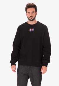 Felpa nera con scollo a barca, caratterizzata da una patch grafica colorata sul petto. Il design include accenti rossi e blu.