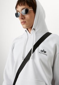 Witte hoodie met trekkoorden, voorzien van een logo op de borst. Een zwarte schouderband en zilveren zonnebril maken de look af.