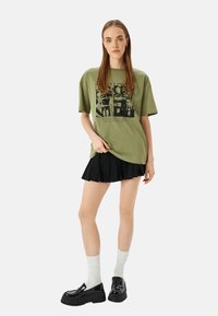Camiseta oversize verde oliva con estampado gráfico negro, combinada con una falda plisada negra y zapatos negros gruesos; modelo lleva calcetines blancos.