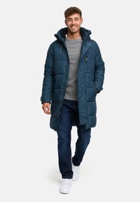 Cămașă puffer teal cu glugă, având un design matlasat și fermoar frontal. Purtați peste un pulover gri, asociată cu blugi închiși la culoare și teniși albi.