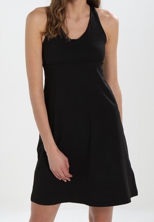 Mujer con vestido negro sin mangas y escote halter, de pie frente a un fondo blanco y liso.