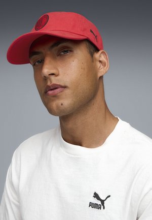 Mężczyzna w czerwonej czapce Puma i białej koszulce Puma, patrzący lekko na bok, na neutralnym szarym tle.