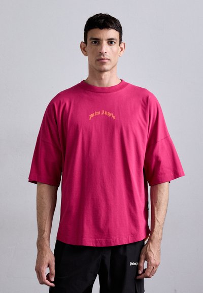 Lyserød bomulds t-shirt med løs pasform og korte ærmer. Har "palm Angels" tekst i guld på brystet. Båret med sorte cargo bukser.