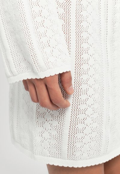 White knitted dress with a textured pattern, featuring openwork sections and a scalloped hem. The sleeve extends with a ribbed edge.