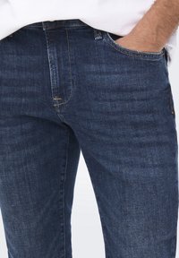 Tummansiniset farkut, joissa on slim fit -malli, hienostunut pinta ja hillitty haalistuminen. Mukana etutaskut ja erottuvat ompeluyksityiskohdat.