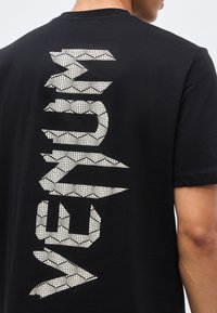 Μαύρο μπλουζάκι με κοντά μανίκια που φοριέται από ένα άτομο, με μεγάλα λευκά γράμματα με σχέδιο που σχηματίζουν τη λέξη "VENUM" κάθετα στην πλάτη.