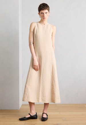 Mujer con el pelo corto, vestida con un vestido midi beige sin mangas y zapatos Mary Jane negros, de pie sobre un suelo de madera frente a una pared lisa y clara.