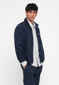 Tamsiai mėlynas lengvas švarkas su stovinčiu apykakle, užtrauktuku, elastinėmis rankogaliais ir šoninėmis kišenėmis, dėvimas ant dryžuoto marškinėlių.