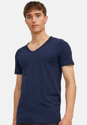 Jeune homme aux cheveux bruns courts portant un t-shirt uni bleu marine à col en V et manches courtes, debout devant un fond uni.