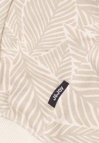 Beige stof met een bladerenpatroon en donkere contouren. Afgebouwde randen met een witte sectie onderaan en een zwart J&Joy-label.