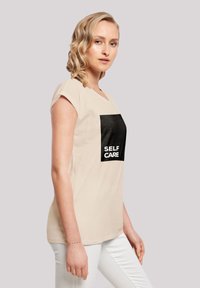 Beige T-Shirt mit kurzen Ärmeln, das ein schwarzes Quadrat mit der Aufschrift "SELF CARE" in weißen Buchstaben zeigt. Glattes Baumwollmaterial.