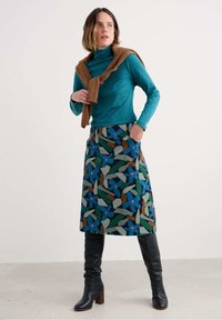 Pulover turcoaz cu guler înalt, pulover maroniu aruncat peste umeri și fustă până la genunchi, cu un model abstract de frunze în nuanțe de albastru, verde și maron. Botine negre până la genunchi.