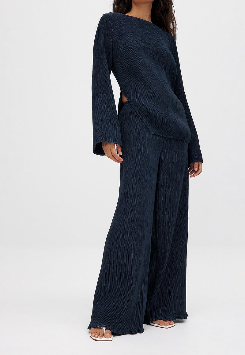 Marineblauw tweedelig outfit met een gestructureerde lange mouw top met een asymmetrische zoom en een wijde broek, beide met een lichte glans.